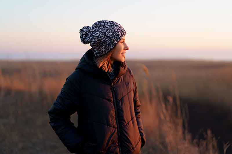 Jeune femme avec bonnet et manteau chaud dans un décor doux de fin d'hiver regarde au loin devant un champ d'herbe