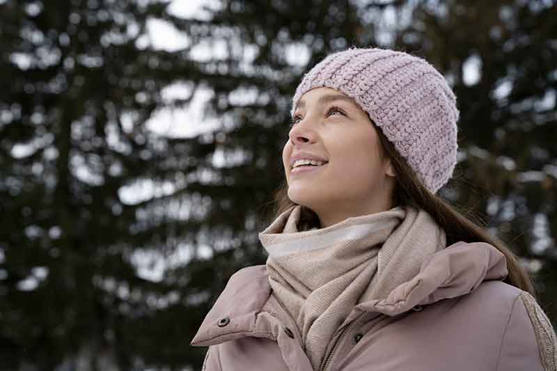 Jeune femme avec bonnet et manteaux roses regarde dans le ciel doux à la fin de l'hiver dans un décor sapineux | Gabriella Chi